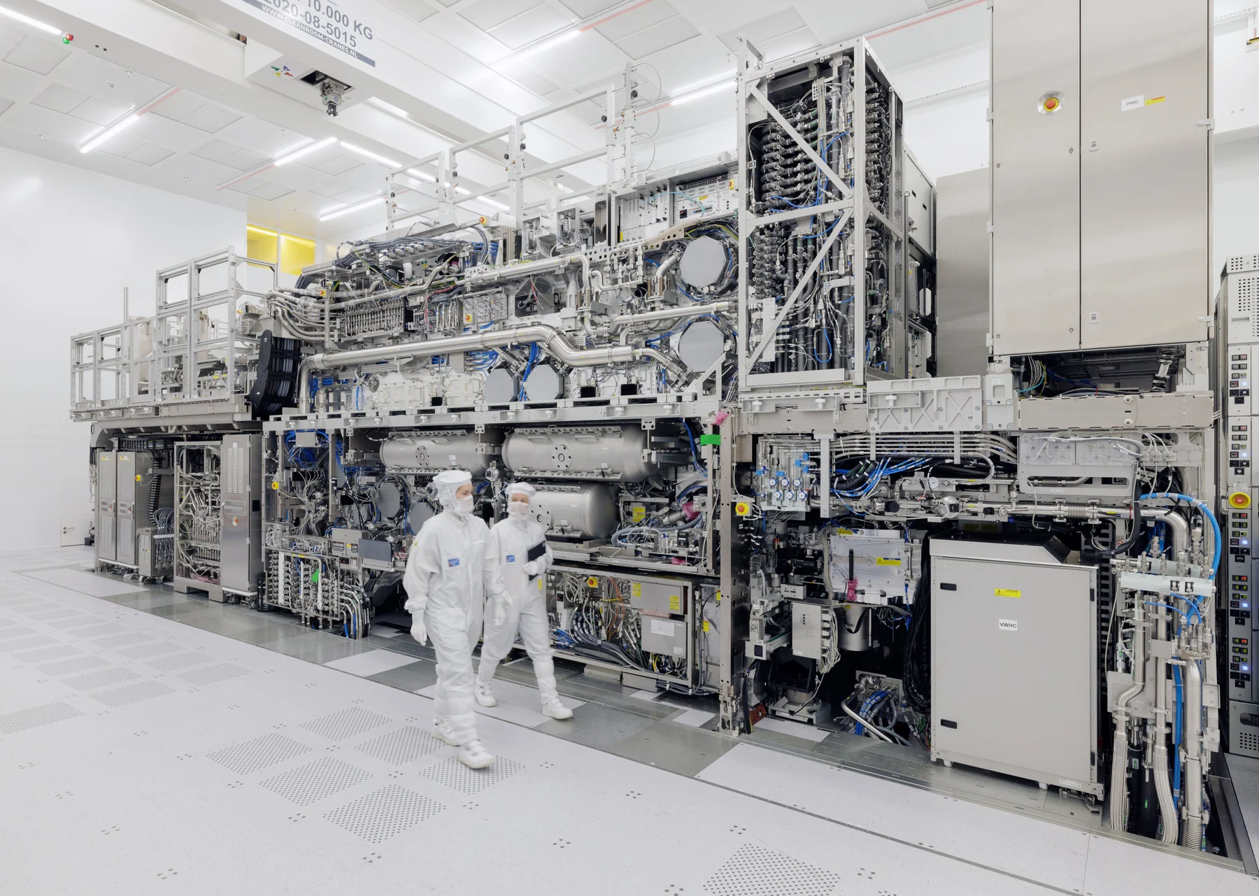 ASML Veldhoven 洁净室内，两名技术人员从一套占据整面空间的 High-NA EUV 光刻系统前走过